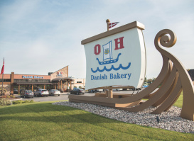 A large Viking ship-shaped sign for O&H Danish Bakery stands on a landscaped lawn outside the bakery’s flagship location. The bakery building, parking area, and a Danish flag are visible in the background on a clear day.