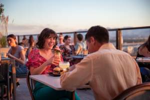 A man and a woman having dinner at a table. The woman is smiling and laughing. The background is a sunset shot over Salt Lake