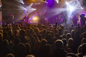 A band performing on stage with bright, colorful lights, while a large crowd watches, with their hands raised.