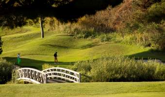 Ben Brown Golf Course at The Ranch at Laguna Beach, featuring a scenic fairway surrounded by rolling green hills, a white arched footbridge crossing a natural area, and golfers playing beneath mature trees in a tranquil canyon setting.