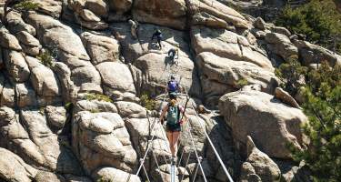 person walking across wire bridge while people ahead are climbing on rock face while clipped into iron rope.