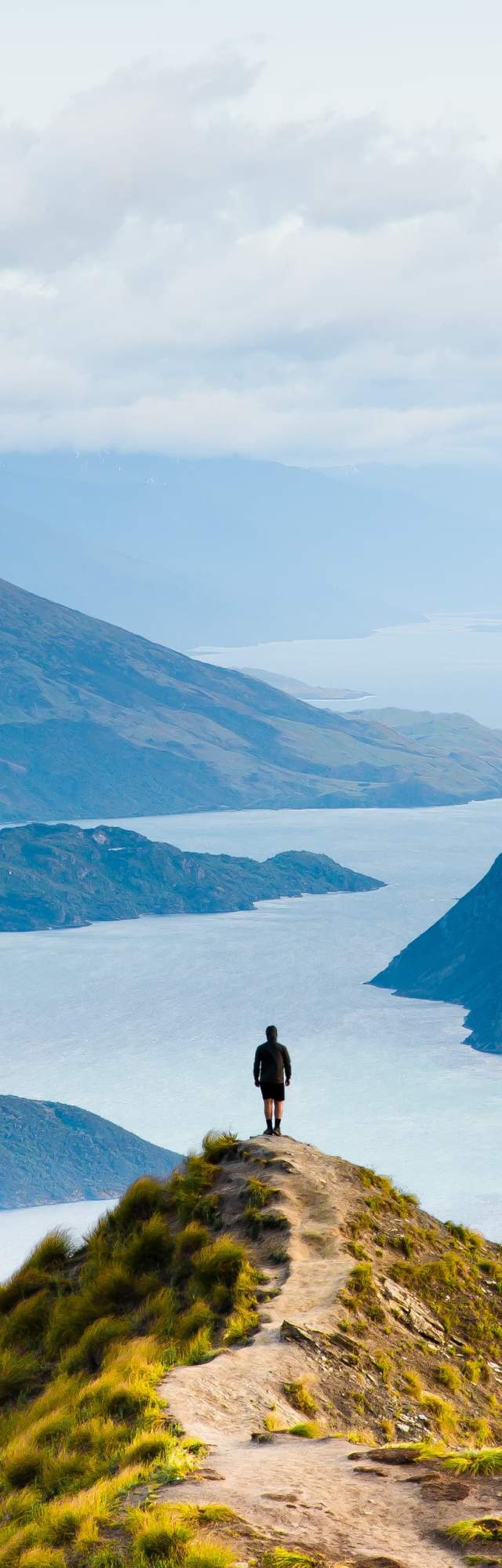 Person standing on cliff overlooking water and mountains.