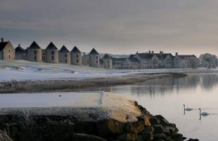 Snow-covered Lough Erne Resort reflected in a calm lake at winter sunrise, with two swans gliding across the water.