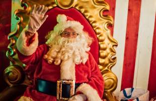 A jolly looking Santa waves as he sits on a plush throne in his grotto