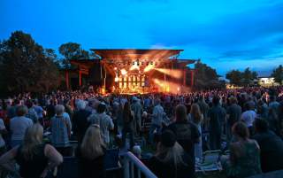 Crowd of people at dusk watching a brightly lit stage with a cool late evening sky