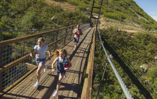 Father and two girls running across a walking path suspension bridge with green mountainside in the background