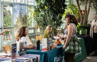 In a greenhouse room with lots of plants a woman is standing in front of colorful vendor tables talking to the people sitting behind them.