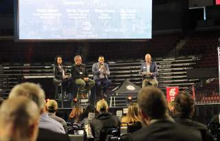 Panel of 2 men and one woman on stage with a moderator asking questions and a presentation on the screen above and behind them