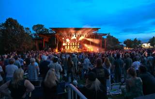 Crowd of people at dusk watching a brightly lit stage with a cool late evening sky