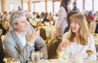 Two well dressed people smiling and talking at a set table with more set tables and people in the background