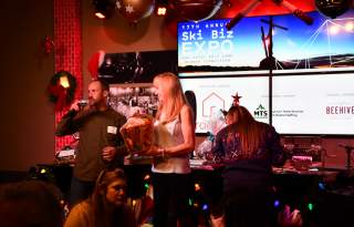 Two people on microphones one with a gift in her hands. The background has TV screens that say 17th annual Ski Biz Expo