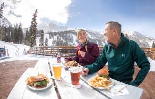 Man and woman in light winter gear sitting at an outdoor table eating with snowy mountains and skis in the background