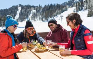 4 people sitting at an outdoor table with food, solitude mountain with snow and a lift in the background