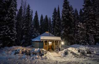 A yurt lit up by lanterns on each side of the door in the snow with large pines in the background