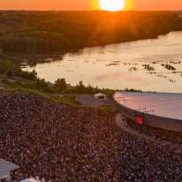 Aerial shot of Lakeview Amphitheater, large crowd in front of amphitheater set on the shores of Onondaga Lake, a fiery orange sunset can be seen in the horizon