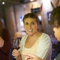 Young Female Smiles in Sweater with Wine Glass in Hand Chatting with Friends