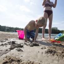 Young Boy in Swimwear Plays in Sand with Pink Bucket as Mom Stands Watching in Background