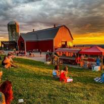 a sunset in back of a red barn with people sitting on the lawn in the foreground