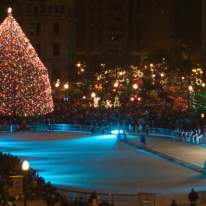 A bright lit up Christmas tree at night time overlooking an empty ice skating rink surrounded by a mass of people.