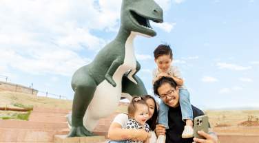 A happy family takes a selfie in front of a large green and white dinosaur statue on a sunny day, with smiles and playful expressions.