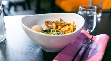 A bowl of ramen on a dark table, featuring a soft-boiled egg, mushrooms, and green onions. Nearby, there is a glass of water and black chopsticks on a pink napkin.