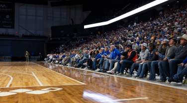 Large crowd seated courtside and in arena stands during a basketball game, with fans watching the action from rows along the hardwood floor under bright indoor lighting.