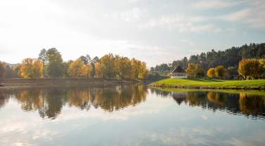 A perfectly still lake in the foreground perfectly reflects trees on the shore in the middle of the image, in fall hues of yellow and green. A small gazebo sits on an island just to the right of the center of the image, also reflected in the water.