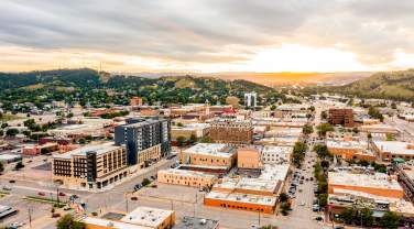 Aerial view of downtown Rapid City at sunset, with downtown streets lined by low-rise buildings, cars, and parking lots. Rolling green hills surround the city, while dramatic clouds stretch across the sky and golden sunlight glows on the horizon.