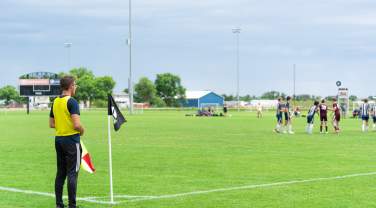 Assistant referee standing at the corner of a green soccer field holding a flag, with players gathered in the distance near midfield under a cloudy sky and stadium lights.
