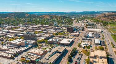 Aerial view of downtown Rapid City with buildings, roads, and parking lots. Surrounding green hills and a clear blue sky create a serene and expansive atmosphere.