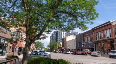 St. Joseph Street in downtown Rapid City with brick buildings, leafy trees and parked cars under a clear blue sky. The scene conveys a vibrant, sunny atmosphere.