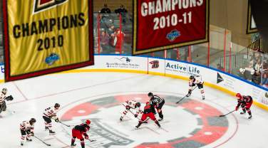 Hockey players from two teams line up for a faceoff while a referee drops the puck, with fans in the stands behind the glass and large championship banners hanging overhead in the arena.