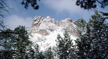 Snow-covered Mount Rushmore is framed by pine trees under a blue sky. The carved faces stand out amid the white, creating a serene winter scene.