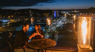 Couple enjoying drinks at a rooftop bar beside a glass fire feature, overlooking the glowing city lights of Rapid City at dusk. The pair sits at a small table, toasting mugs while the skyline and Black Hills foothills stretch into the evening background.