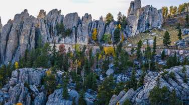 Jagged granite spires tower over a forested landscape with scattered autumn foliage. The scene is serene and dramatic under a partly cloudy sky.