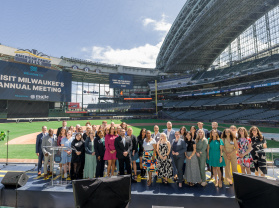 Group photo of attendees at Visit Milwaukee's Annual Meeting, posed on a stage at American Family Field (Milwaukee Brewers stadium) with stadium seating and banners visible in the background.
