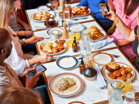Overhead photo of a group of people gathered around a restaurant table filled with breakfast and brunch dishes. Plates include eggs and toast, bacon, a burger with fries, a sauced entrée, and other shared items, along with drinks like orange juice and water. Several people are holding phones and taking photos of the food.