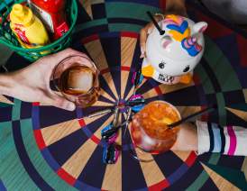Hands cheersing cocktails over a dart board shaped table.