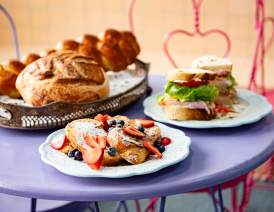 Food from the Denison Bakery is displayed on a small purple cafe table. The front plate has some triangular slices of French toast with powdered sugar and mixed berries on top. The back right plate is a sandwich filled with meat, tomato, and lettuce. The tray to the left displays some of their handmade sourdough breads.