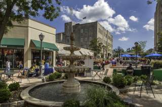 Suburban Square located in Ardmore, PA. The fountain is located in the middle of the shopping district. In the background is the storefront of Barbour. Vendors are seated at tables talking to customers. Many individuals are seen walking around enjoying the warm weather or seated outside for dining at the tables with blue umbrellas.