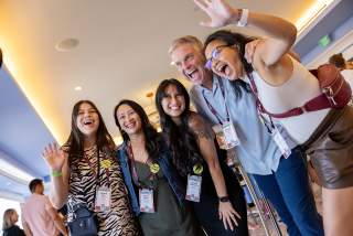 Group of 4 women and 1 man smiling and posing for the camera, angled up towards them from below.