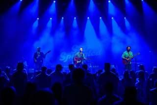 Band playing on a stage with shadows of fans in the foreground. The whole image is lit by blue lights from above.