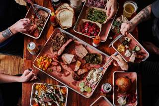 Overhead image of a table full of plates of smoked barbecue and hands reaching into the frame holding utensils and grabbing food.