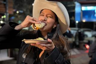 Woman in all black and a cowboy hat joyfully biting into a full taco.