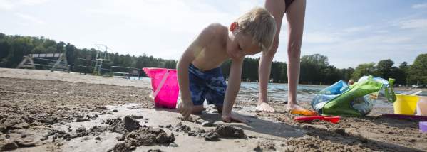 Young Boy in Swimwear Plays in Sand with Pink Bucket as Mom Stands Watching in Background