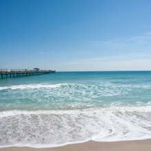 Ocean waves gently crashing onto the Lauderdale-By-The-Sea beach with a long wooden pier extending into the clear blue water under a sunny sky.