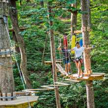 Two people navigate treetops at Treetop Quest Gwinnett, surrounded by green foliage and wooden platforms.