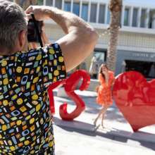 Man in patterned shirt taking a photograph of a woman in an orange dress posing among large red heart sculptures outdoors.