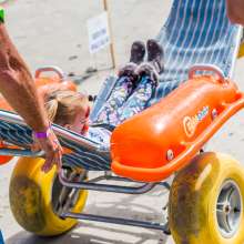 A young girl is pushed on the beach in a large-wheeled wheelchair