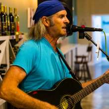 A man in a light blue shirt and dark blue hat strums a guitar and sings into a microphone.